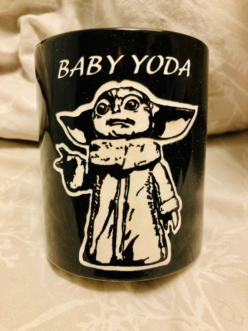 Pode incluir: Caneca de cer&acirc;mica preta com uma imagem branca do Baby Yoda de Star Wars. A caneca tem o texto "BABY YODA" impresso nela.