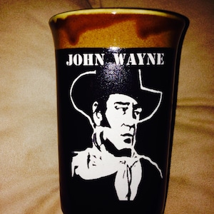 Puede incluir: Una taza de cerámica negra con un borde marrón y una imagen blanca de John Wayne con un sombrero de vaquero. La taza tiene el texto "John Wayne" impreso en ella.