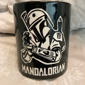 May include: Black ceramic mug featuring a white graphic of a Mandalorian character with a blaster. The word "MANDALORIAN" is printed in white below the graphic. The mug has a glossy finish.