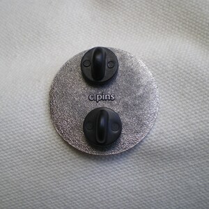 Puede incluir: Un broche redondo de color plateado con dos broches de metal negro y el texto "c.pins" impreso en la parte posterior.