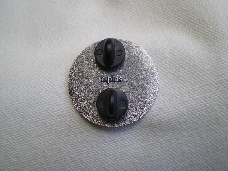 Puede incluir: Un broche redondo de color plateado con dos broches de metal negro y el texto "c.pins" impreso en la parte posterior.