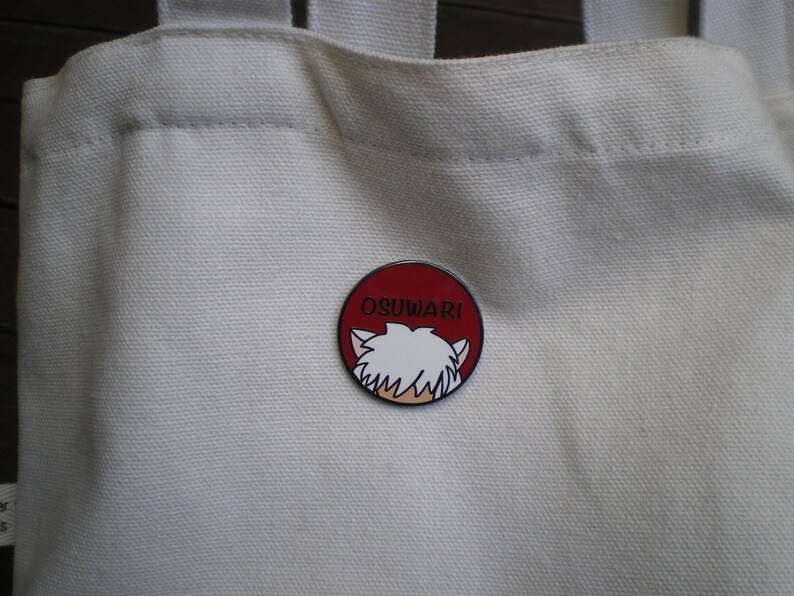 Puede incluir: Bolsa de lona blanca con un pin de esmalte rojo y blanco con un personaje de dibujos animados con cabello blanco y el texto "OSUWARI".
