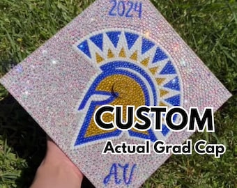 May include: A white graduation cap covered in rhinestones with a blue and gold Spartan helmet design. The cap has the year "2024" and the word "CUSTOM" printed on it. The text "Actual Grad Cap" is also printed on the cap.