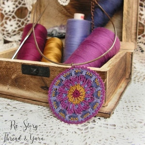 May include: A crocheted mandala pendant in shades of purple, pink, and yellow hangs from a copper chain. The pendant is displayed in a wooden box with spools of thread in various colors.
