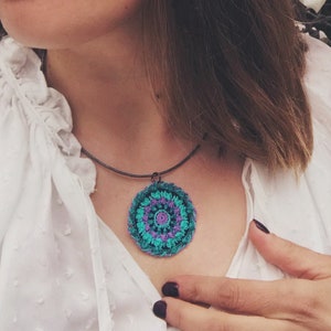 Op de afbeelding: Een vrouw draagt een wit shirt met een gehaakte hangerketting met een turquoise, paars en roze mandala-ontwerp. De ketting is aan een zwart koord bevestigd. De tekst "Re-Story Thread & Yarn" is zichtbaar in de rechterbenedenhoek van de afbeelding.