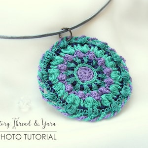 Op de afbeelding: Een gehaakte ketting met een hanger in teal, paars en donkerblauw met een mandala-ontwerp. De hanger is bevestigd aan een zilveren ketting.