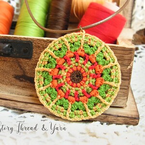 May include: A crocheted pendant with a circular design in shades of green, orange, and red. The pendant is hanging from a brown leather cord and is resting on a wooden box.