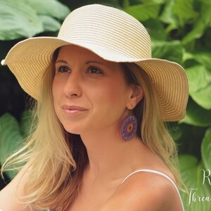 May include: A woman wearing a wide-brimmed straw hat and a white tank top. She is wearing large, round, crocheted earrings with purple, yellow, and orange accents. The background is a lush green garden.