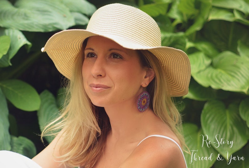 May include: A woman wearing a wide-brimmed straw hat and a white tank top. She is wearing large, round, crocheted earrings with purple, yellow, and orange accents. The background is a lush green garden.