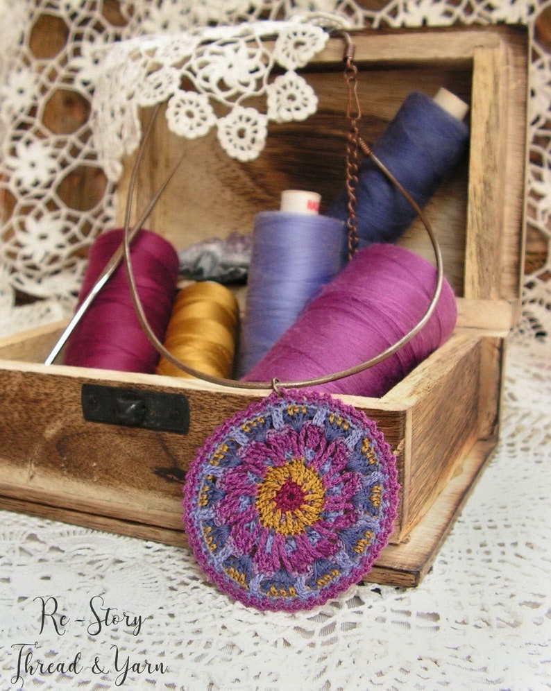 May include: A crocheted mandala pendant in shades of purple, pink, and yellow hangs from a copper chain. The pendant is displayed in a wooden box with spools of thread in various colors.