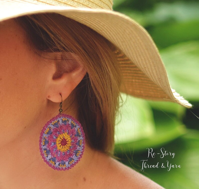 May include: A pair of crocheted earrings with a pink, purple, and yellow mandala design. The earrings are hanging from a woman's ear. The text "Re-Story Thread & Yarn" is visible in the bottom right corner of the image.