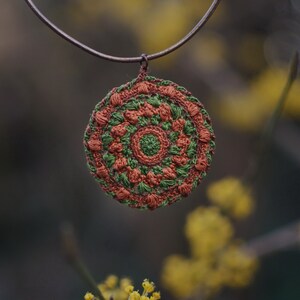 Op de afbeelding: Een gehaakte mandala-hanger in tinten bruin en groen hangt aan een bruin koordsnoer. De hanger is rond met een centrale cirkel en stralende spaken. De achtergrond is wazig en toont gele bloemen.