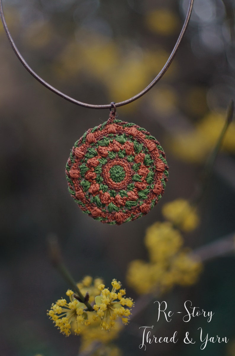 Op de afbeelding: Een gehaakte mandala-hanger in tinten bruin en groen hangt aan een bruin koordsnoer. De hanger is rond met een centrale cirkel en stralende spaken. De achtergrond is wazig en toont gele bloemen.