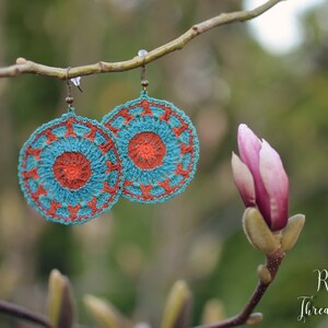 May include: A pair of crocheted earrings with a teal and orange color scheme. The earrings are hanging from a branch with a pink flower bud in the background. The text "Re-Story Thread & Yarn" is visible in the bottom right corner of the image.