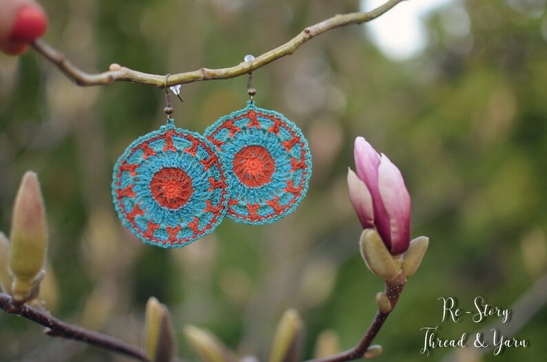 May include: A pair of crocheted earrings with a teal and orange color scheme. The earrings are hanging from a branch with a pink flower bud in the background. The text "Re-Story Thread & Yarn" is visible in the bottom right corner of the image.