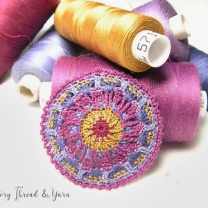 May include: A close-up of a crocheted doily with a multi-colored design. The doily is made with shades of pink, purple, and gold thread. The doily is surrounded by spools of thread in various colors.