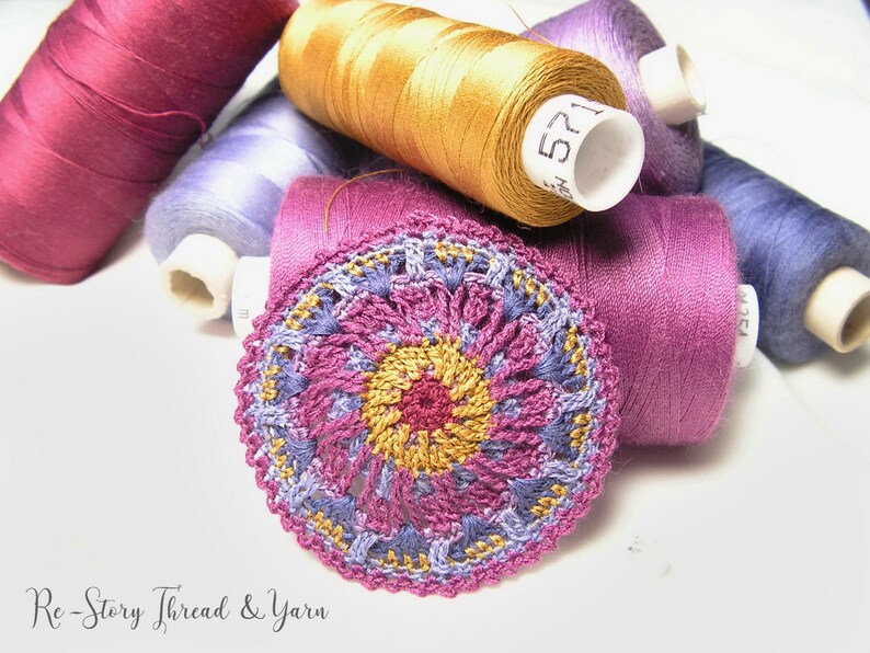 May include: A close-up of a crocheted doily with a multi-colored design. The doily is made with shades of pink, purple, and gold thread. The doily is surrounded by spools of thread in various colors.
