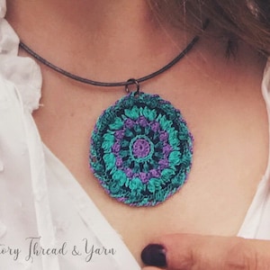 Op de afbeelding: Een gehaakte mandala-hangerketting met een turquoise, paarse en zwarte kleurstelling. De ketting is aan een zwart koord bevestigd en wordt door iemand gedragen.