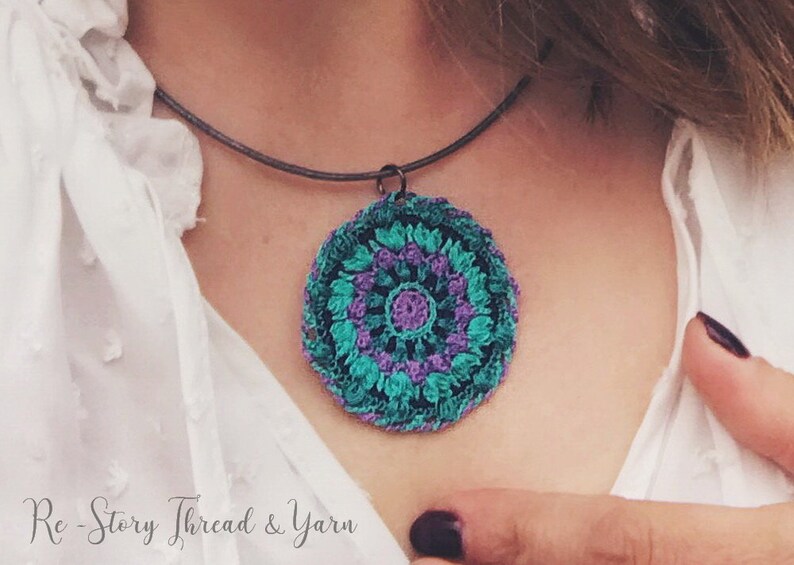 Op de afbeelding: Een gehaakte mandala-hangerketting met een turquoise, paarse en zwarte kleurstelling. De ketting is aan een zwart koord bevestigd en wordt door iemand gedragen.