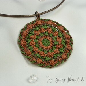 Op de afbeelding: Een gehaakte ketting met een hanger in bruin en groen, met een mandala-ontwerp. De ketting is gemaakt van een bruin leren koord.