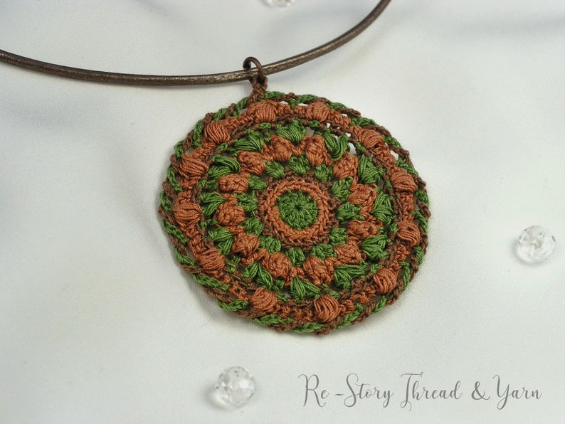 Op de afbeelding: Een gehaakte ketting met een hanger in bruin en groen, met een mandala-ontwerp. De ketting is gemaakt van een bruin leren koord.