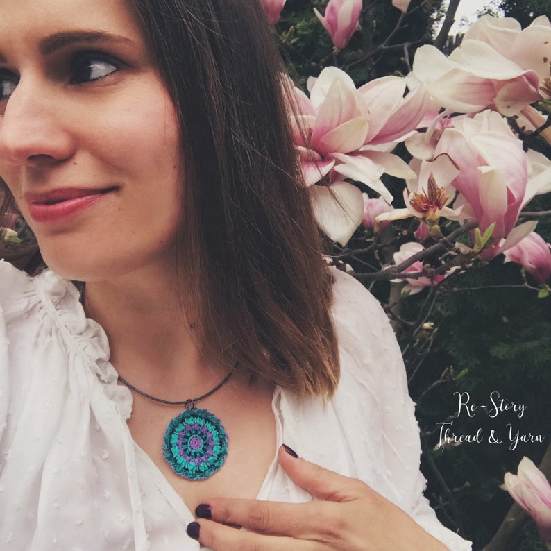 Op de afbeelding: Een vrouw draagt een wit shirt met een gehaakte hangerketting met een turquoise, paars en roze mandala-ontwerp. De ketting is aan een zwart koord bevestigd. De tekst "Re-Story Thread & Yarn" is zichtbaar in de rechterbenedenhoek van de afbeelding.