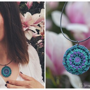 Op de afbeelding: Een gehaakte mandala-hangerketting met een turquoise, paarse en witte kleurstelling. De ketting hangt aan een zwart koord en is gefotografeerd tegen een wazige achtergrond van roze bloemen.