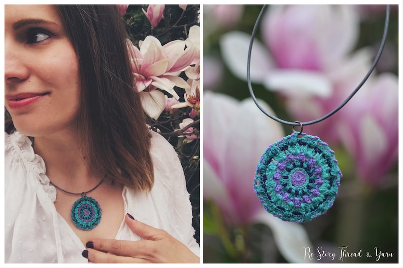 Op de afbeelding: Een gehaakte mandala-hangerketting met een turquoise, paarse en witte kleurstelling. De ketting hangt aan een zwart koord en is gefotografeerd tegen een wazige achtergrond van roze bloemen.