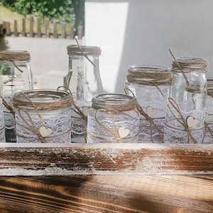 Könnte beinhalten: Ein rustikales Holztablett mit durchsichtigen Glasbehältern, die mit Spitze und Schnur verziert sind. Jeder Behälter hat ein kleines Holzhäuschen angebracht. Die Behälter eignen sich perfekt zum Aufbewahren von Kerzen oder anderen Dekorationsartikeln.