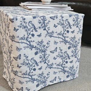 Può includere: Un pouf in tessuto bianco e blu con un motivo floreale con uccelli e farfalle. Il pouf è seduto su un tappeto grigio davanti a un divano in pelle nera.