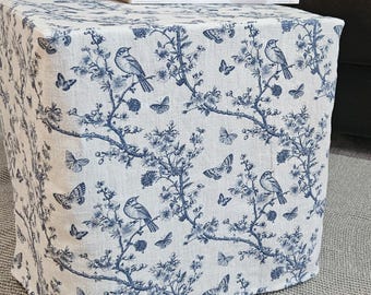 Puede incluir: Un otomano de tela blanco y azul con un diseño floral con pájaros y mariposas. El otomano está sentado en una alfombra gris frente a un sofá de cuero negro.