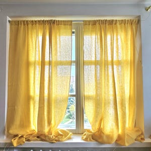 Puede incluir: Cortinas de lino amarillas colgadas frente a una ventana. Las cortinas están recogidas en la parte superior y caen, con la luz del sol filtrándose a través de la tela. El marco de la ventana es blanco y las cortinas son de un amarillo cálido y soleado.