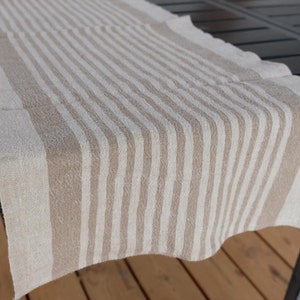 May include: A beige and white striped linen table runner. The runner is made of a soft, textured fabric and has a subtle sheen.