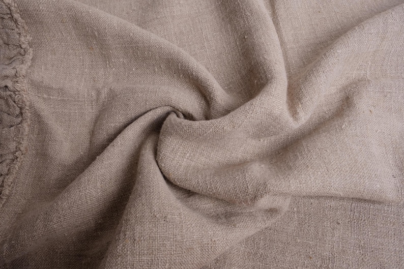 May include: A close-up of a natural linen fabric with a textured, woven surface. The fabric is folded and draped, creating soft folds and wrinkles.