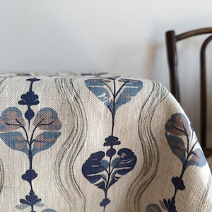 Può includere: Un tovaglia di lino beige con un motivo floreale blu e marrone. La tovaglia ha un design ondulato e presenta motivi floreali stilizzati.