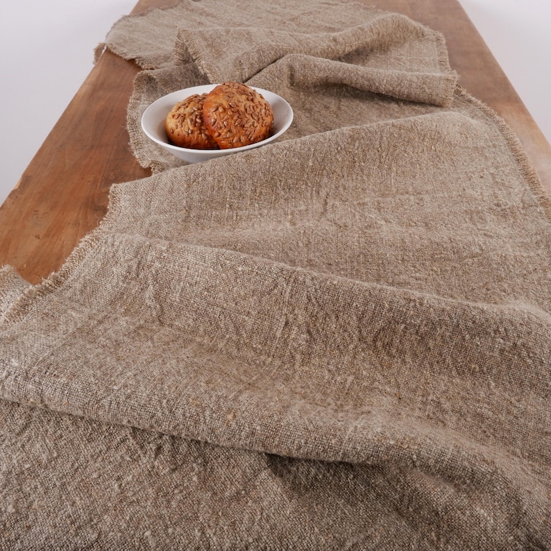Puede incluir: Un camino de mesa de lino beige con borde deshilachado, sobre una superficie de madera. Un cuenco blanco peque&ntilde;o contiene dos panecillos redondos con semillas. La tela natural a&ntilde;ade calidez y textura.