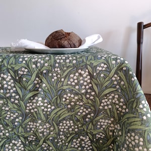 Può includere: Una tovaglia verde con un motivo floreale bianco. La tovaglia è in lino e ha una leggera lucentezza. Un pane è appoggiato su un piatto bianco sopra la tovaglia.