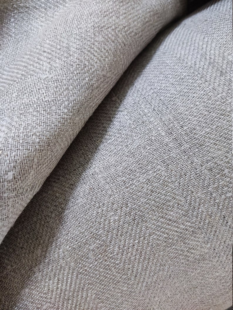 May include: Close-up of a light grey fabric with a subtle herringbone pattern. The fabric is woven with a textured, slightly rough surface.