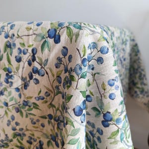 May include: A white linen tablecloth with a blue and green blueberry print. The tablecloth is made of natural linen and has a soft, textured feel.