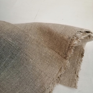 Puede incluir: Primer plano de un trozo de tela de lino natural con un borde deshilachado. La tela es de color marr&oacute;n claro y tiene una textura rugosa.