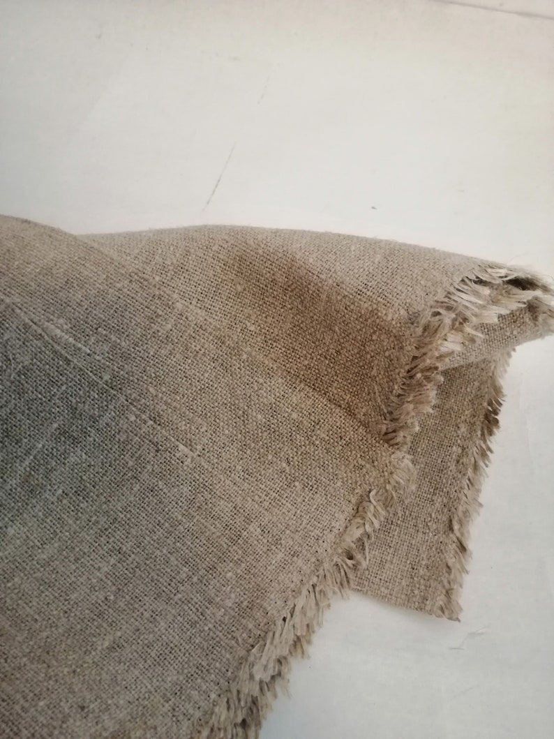 Puede incluir: Primer plano de un trozo de tela de lino natural con un borde deshilachado. La tela es de color marr&oacute;n claro y tiene una textura rugosa.