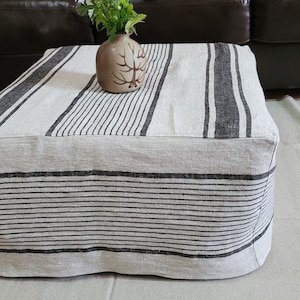 May include: A square, beige ottoman with black vertical stripes. A small, brown vase with a tree design holds blue and white flowers. The ottoman is on a light beige rug, with a dark brown leather couch in the background.