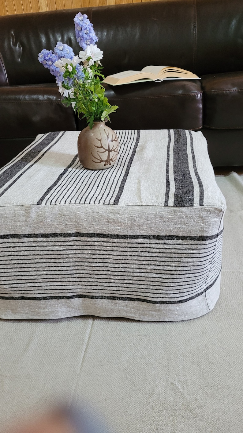 May include: A square, beige ottoman with black vertical stripes. A small, brown vase with a tree design holds blue and white flowers. The ottoman is on a light beige rug, with a dark brown leather couch in the background.