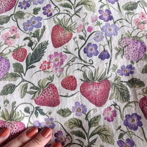 May include: A fabric with a repeating pattern of strawberries, flowers, and leaves. The strawberries are red and purple, the flowers are purple and pink, and the leaves are green. The background is a light cream color.
