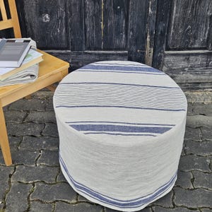 May include: A circular ottoman with a light grey fabric cover, adorned with horizontal blue stripes and a blue border. A wooden chair and books are visible in the background. The ottoman is a stylish piece of furniture.