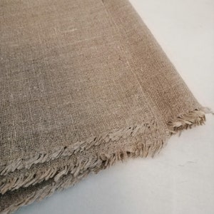 Puede incluir: Primer plano de una tela de lino natural con un borde sin terminar. La tela es de color beige claro y tiene una superficie ligeramente texturizada.