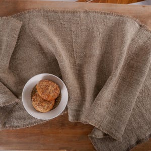 Puede incluir: Un camino de mesa de lino beige con borde deshilachado se extiende sobre una superficie de madera. Un cuenco blanco con borde dorado contiene dos panecillos redondos con semillas, creando una escena acogedora.