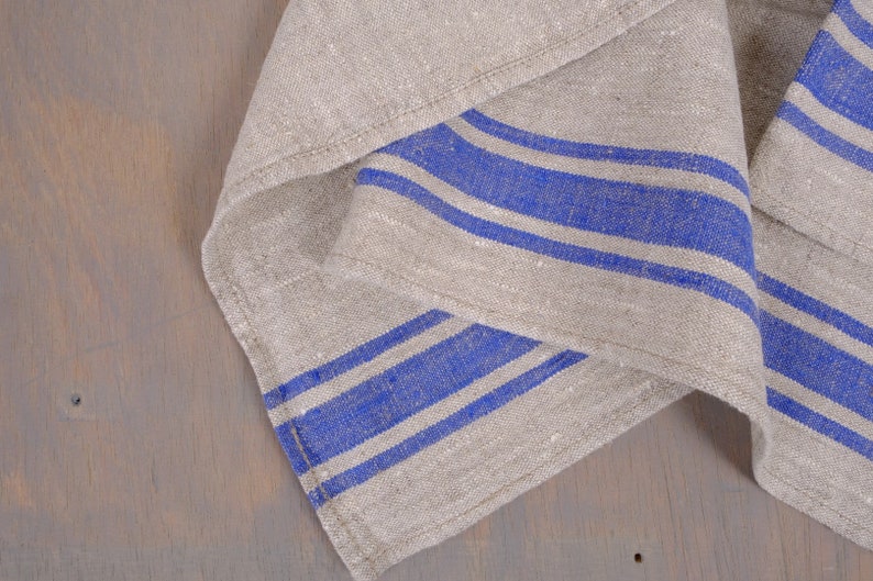 May include: A folded linen kitchen towel with blue stripes. The towel is made of natural linen fabric and has a rustic look.