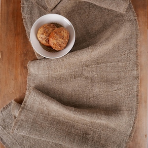 Puede incluir: Un camino de mesa de lino marr&oacute;n texturizado con un borde deshilachado. Un cuenco blanco con un borde decorativo contiene dos panecillos con semillas. El camino de mesa est&aacute; sobre una superficie de madera.