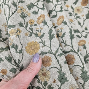May include: A close-up of a beige fabric with a floral pattern. The fabric features small yellow flowers with green leaves on a beige background.